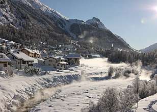 Winterliche Dorfansicht von Pontresina mit verschneiten Häusern, dampfendem Bach und imposantem Bergpanorama bei strahlend blauem Himmel