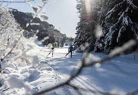 Two cross-country skiers trace their tracks through the snow-covered trail in the Engadin, with a historic mountain hotel in the background among snow-laden fir trees