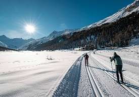 Zwei Langläufer auf einer breiten Loipe im Engadin mit weitem Talblick und strahlender Wintersonne.